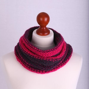 Könnte beinhalten: Ein gestrickter Rundschal in Pink- und Dunkelvioletttönen. Der Schal wird auf einem weißen Schaufensterpuppenkopf mit einem Holzhalspfosten präsentiert. Der Schal ist aus dickem Garn gefertigt und hat eine gerippte Textur.