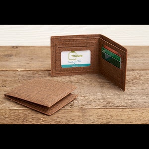 Puede incluir: Cartera marrón con apariencia de tela texturizada, mostrada abierta y cerrada sobre una superficie de madera. La cartera abierta revela ranuras para tarjetas y una ventana transparente con el logotipo "Tofuture". La cartera cerrada está colocada junto a la abierta.