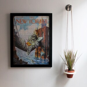 Könnte beinhalten: Ein gerahmtes Cover des New Yorker Magazins mit einem Puzzlebild der Freiheitsstatue, die sich in einer Hängematte über einem Gewässer entspannt. Eine Hängepflanze mit grünen und roten Blättern hängt an einem Haken an der Wand.
