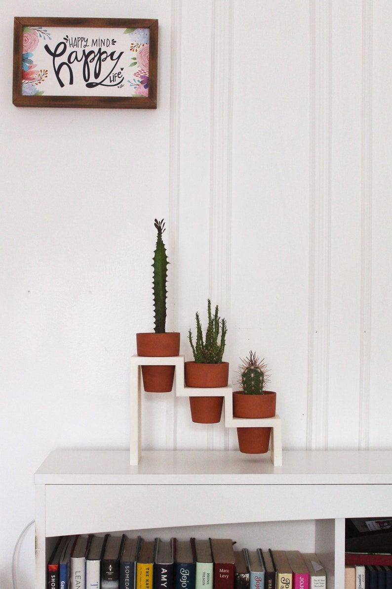 May include: White wooden plant stand with three tiers, featuring three potted succulents. The stand is on a white shelf with books underneath. A framed print with the text "Happy Mind Happy Life" is on the wall above the stand.