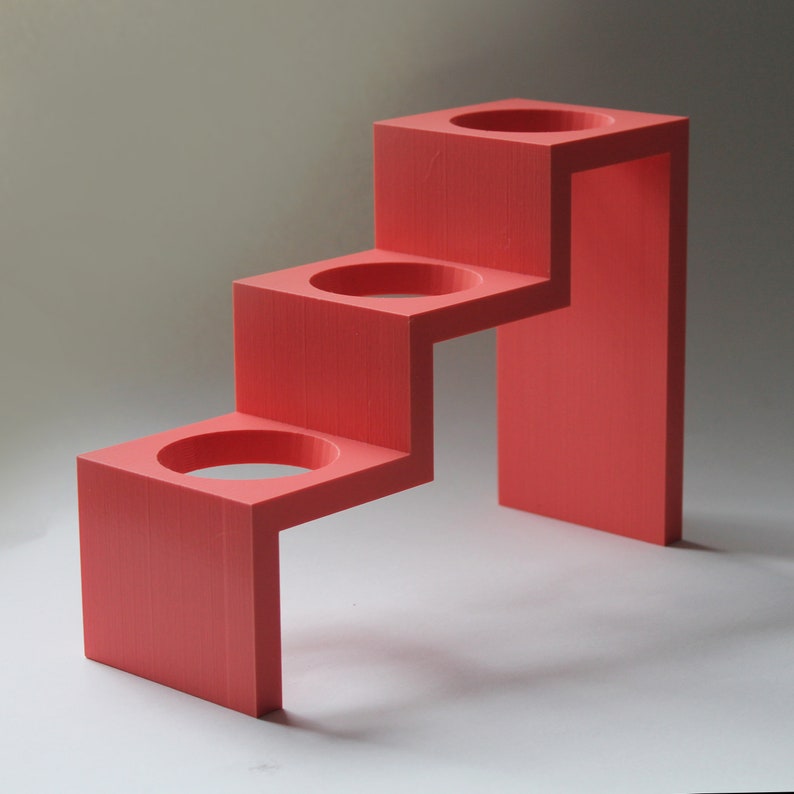 May include: A three-tiered, coral-colored, plastic stand with three circular cutouts. The stand is designed to hold small items, such as candles or toiletries.