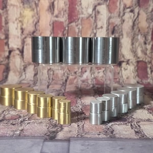 May include: An assortment of metal cylinders in various sizes and finishes. The cylinders are arranged in rows, with some appearing to be brass and others silver-colored. The background is a brick wall.