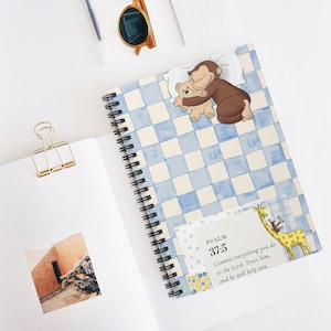 May include: Spiral-bound notebook with a blue and white checkered pattern. The cover features a cartoon monkey hugging a teddy bear. The notebook is open, revealing blank pages and a gold paper clip. Sunglasses and a notepad are also visible.