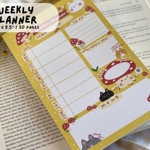 May include: A yellow weekly planner with 50 pages. The planner has a mushroom and animal theme. The cover features a mushroom with a white cat on top, a mushroom with a black cat on top, and a mushroom with a gray cat on top. The planner has a section for "To Do", "Brain", "Memo", and "Dump".