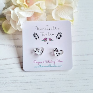 May include: Heart-shaped earrings featuring musical notation, presented on a card with the text "The Reversible Robin". The card also states "Perspex & Sterling Silver" and a website address. The earrings are on a white wooden surface.
