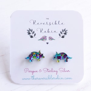 May include: A pair of iridescent blue and purple dinosaur stud earrings with silver posts. The earrings are made of Perspex and sterling silver. The earrings are displayed on a white card with the text "The Reversible Robin" and "www.thereversiblerobin.com".