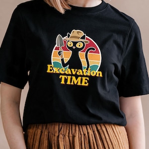 Può includere: Una t-shirt nera a maniche corte con la grafica di un gatto nero che indossa un cappello fedora beige e tiene in mano una piccola paletta da giardinaggio. Dietro il gatto c'è un disegno circolare di un tramonto nei toni arancio, giallo e ottanio.