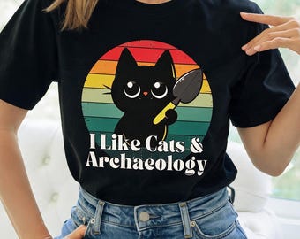 May include: A black short-sleeved t-shirt featuring a graphic of a black cat holding a small trowel inside a circular sunset background with horizontal stripes in red, orange, and green. The text "I Like Cats & Archaeology" is printed in white.