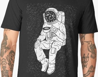 Può includere: Una maglietta nera con un'illustrazione bianca di un astronauta che beve da una tazza nello spazio. L'astronauta indossa un casco con un quadrante dell'orologio su di esso. Lo sfondo è un cielo stellato bianco e nero.