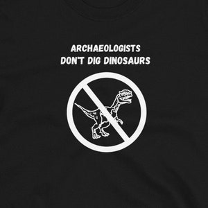 Peut inclure: T-shirt noir avec du texte blanc qui dit "Les archéologues ne creusent pas les dinosaures" et un cercle blanc avec une ligne diagonale qui le traverse et un dinosaure à l'intérieur.