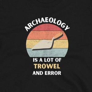 Peut inclure: T-shirt noir avec un graphique de style vintage qui dit "L'archéologie, c'est beaucoup de truelle et d'erreurs". Le graphique représente une truelle et un coucher de soleil.