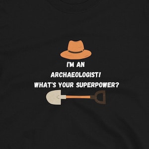 Op de afbeelding: Zwart T-shirt met witte tekst die luidt: "Ik ben een archeoloog! Wat is jouw superkracht?" Een bruine fedora en een schop zijn afgebeeld boven de tekst.