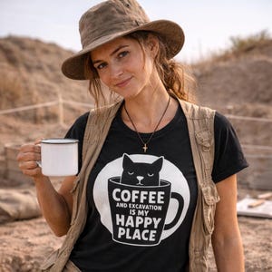 Peut inclure: T-shirt noir avec un motif blanc d'un chat dans une tasse à café. Le texte sur le t-shirt se lit « Coffee and Excavation is my Happy Place ». La personne porte un chapeau et un gilet marron et tient une tasse blanche.