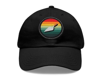 Puede incluir: Gorra de béisbol negra con un parche circular con un diseño de llana sobre un fondo de puesta de sol retro con rayas rojas, naranjas, amarillas y verdes. La llana es blanca con un mango marrón. El parche está delineado con una línea de puntos blanca.