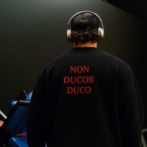 Pode incluir: Camisola preta com o texto vermelho "NON DUCOR DUCO" nas costas. A pessoa está a usar auscultadores dourados e um boné de camionista preto. O fundo é um ambiente escuro de ginásio.