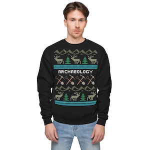 Peut inclure: Sweat-shirt noir avec un motif de pull de Noël représentant des rennes, des sapins et les mots "ARCHAEOLOGY". Le motif comprend également des pioches et des pelles croisées.
