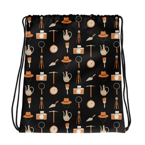 May include: A black drawstring bag with a repeating pattern of orange and brown illustrations. The illustrations include a compass, a camera, a trowel, a teapot, a paintbrush, a magnifying glass, and a hat.