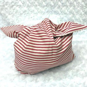 May include: A fabric bag tied in a knot at the top, featuring a pattern of thin horizontal red stripes on an off-white background. The bag has two extended fabric ends that form a knot, creating a structured shape with visible folds and seams.