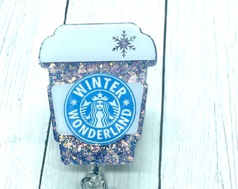 May include: A sparkly, glittery, light blue and white coffee cup with a snowflake on top. The cup has a blue circle with the text "Winter Wonderland" and a mermaid logo inside. The cup is attached to a clear plastic badge holder with a silver clip and a sparkly, glittery, light blue and white round charm.