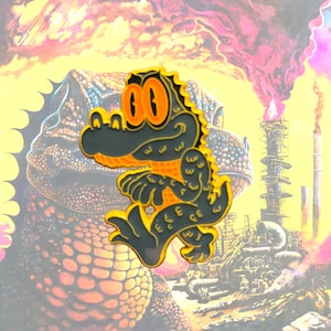 May include: A dark gray and bright yellow enamel pin featuring a cartoon alligator with large orange eyes. The alligator is depicted in a dynamic pose with a textured body, sharp teeth, and a curved tail, outlined in a bold yellow metal border.