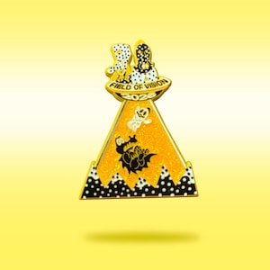 May include: A gold-toned metal enamel pin shaped like a triangular UFO beam. The top features two figures in a saucer labeled FIELD OF VISION 2026. The orange glitter beam contains a small ghost and a black alligator graphic over a polka dot base.