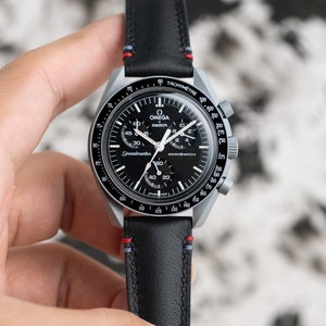 Peut inclure: Une montre-bracelet chronographe Omega Speedmaster Moonwatch argentée et noire avec un bracelet en cuir noir et des coutures rouges et bleues. Le cadran de la montre comporte une échelle tachymétrique et les mots "Speedmaster" et "Moonwatch" imprimés dessus.
