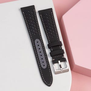 May include: Two-piece watch strap in dark gray canvas with white stitching. The strap features multiple ventilation holes and a gray leather-like backing. A silver-colored buckle is attached to one end.