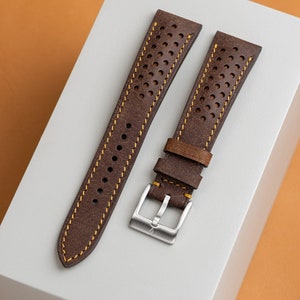 May include: Two-piece brown leather watch strap with yellow stitching and perforated holes. The strap has a silver buckle. The leather has a distressed look. The watch strap is displayed on a white surface with an orange background.