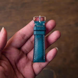 May include: A teal blue leather watch strap with white stitching and a silver buckle.