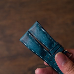 May include: A teal blue leather watch strap with white stitching. The strap is folded in half and held by a hand.