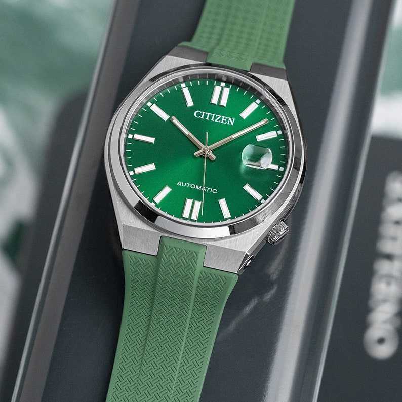 Puede incluir: Un reloj autom&aacute;tico Citizen plateado con una esfera verde y una correa de silicona verde. La esfera del reloj tiene marcadores de hora en n&uacute;meros romanos blancos y una ventana de fecha en la posici&oacute;n de las 3 en punto.