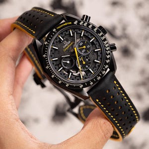 Puede incluir: Un reloj de pulsera cronógrafo negro con una correa de cuero amarilla y negra. La esfera del reloj tiene un diseño esqueletizado y presenta una escala de taquímetro alrededor del borde exterior. La esfera del reloj también incluye el texto "Ω OMEGA Speedmaster".