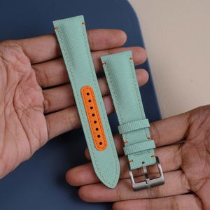 Pode incluir: Pulseira de relógio de duas peças na cor verde-água com costuras e detalhes em laranja. A pulseira apresenta uma fivela de aço inoxidável e uma peça de destaque laranja com vários orifícios para ajuste. O material parece ser lona texturizada.