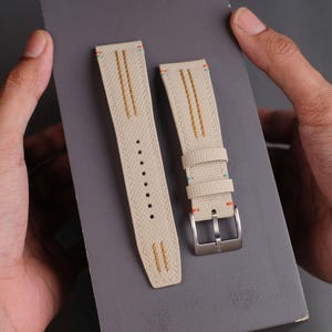 May include: Beige canvas watch straps with orange, teal, and gold stitching. The straps have a silver buckle and are displayed on a gray surface. The watch straps are new and unused.