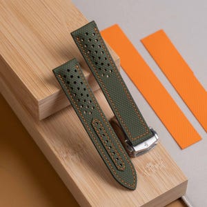 Puede incluir: Una correa de reloj de color verde oliva con textura perforada y costuras de contraste en color naranja brillante. La correa incluye un cierre desplegable de metal plateado y se acompaña de dos inserciones naranjas texturizadas, planas y rectangulares.