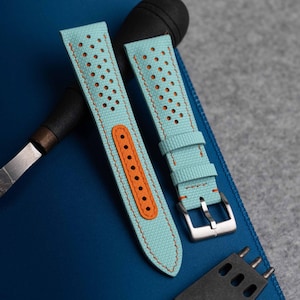 May include: A light blue watch strap featuring a textured surface with perforated holes, orange stitching, and an orange leather lining around the buckle holes. The strap includes a silver-toned metal buckle and two matching light blue keepers.