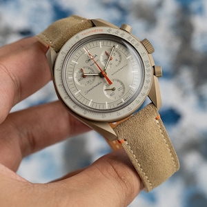 May include: A beige and orange Omega Speedmaster Moonswatch with a tachymeter bezel and a beige suede strap.