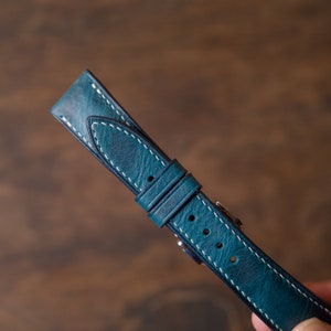 May include: A teal blue leather watch strap with white stitching. The strap has a buckle and is made of high-quality leather.