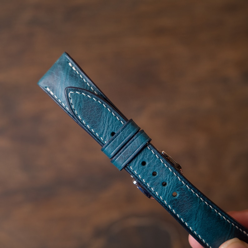 May include: A teal blue leather watch strap with white stitching. The strap has a buckle and is made of high-quality leather.