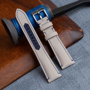 Può includere: Un cinturino per orologio beige con una superficie in tessuto testurizzato e cuciture blu a contrasto. Il cinturino include un rinforzo in pelle blu scuro con fori punzonati e una fibbia in metallo color argento all'estremità.