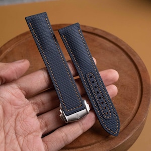 May include: A navy blue watch strap with a textured fabric surface and contrasting orange stitching. The strap features a silver-toned metal deployment clasp with a small engraved logo at the base and multiple adjustment holes for sizing.