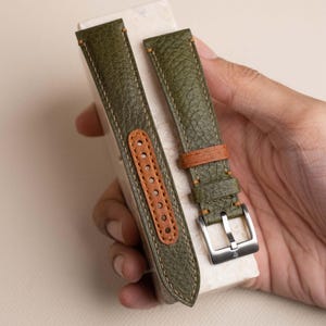 May include: A two-piece olive green textured leather watch strap featuring tan stitching and a tan leather lining with punched holes. The strap includes a silver-toned metal buckle and a tan leather keeper loop, showcasing a rugged finish.