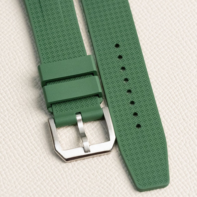 Puede incluir: Una correa de reloj de caucho verde con una hebilla plateada. La correa tiene un patr&oacute;n texturizado.