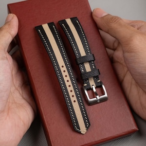May include: Two black and tan watch straps with white stitching and a silver buckle. The straps are held over a red surface. The watch straps have a striped design.