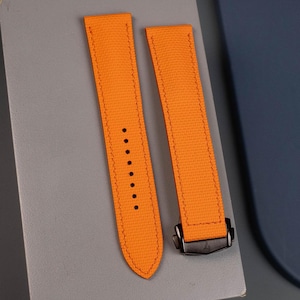 May include: Bright orange watch strap with black clasp. The strap is made of a textured material and features stitching along the edges and several holes for adjustment. The clasp is a dark, matte finish.