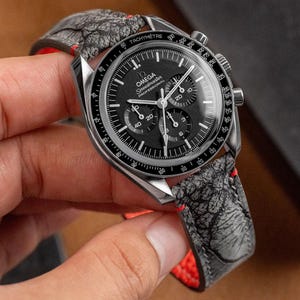 Puede incluir: Un reloj de pulsera cronógrafo negro y plateado con una correa de cuero negro con un diseño estampado en gris y blanco. La esfera del reloj tiene una escala de taquímetro y las palabras "Omega Speedmaster Professional" impresas en ella.