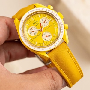 May include: A yellow Omega Speedmaster Moonwatch chronograph wristwatch with a yellow leather band. The watch face has a tachymeter scale, a white dial with black markings, and a red second hand.