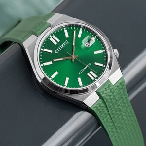 Puede incluir: Un reloj autom&aacute;tico Citizen plateado con una esfera verde y una correa de silicona verde. El reloj tiene marcadores de hora y manecillas blancas, y una ventana de fecha en la posici&oacute;n de las 3 en punto.