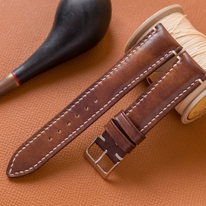 May include: Two brown leather watch straps with white stitching. The straps are laid out on a brown leather surface. One strap is wrapped around a spool of white thread.