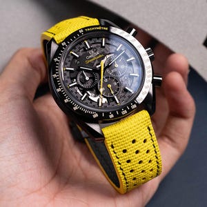 May include: Black Omega Speedmaster watch with a yellow strap. The watch face is black with white and yellow accents, including the word "Speedmaster". The yellow strap has a perforated design.